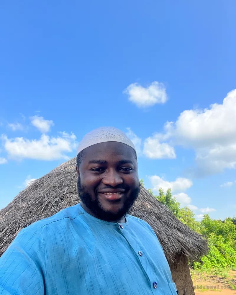 Abdulsamii smiling outdoors under a blue sky beside a thatched hut